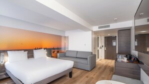 A modern hotel room with a large bed, a grey sofa, a small table, and a wall mural. - Novotel Deauville Plage (Deauville)