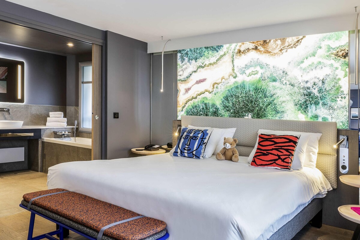 A modern hotel room with a large bed, a bench, a bedside table, a nightstand, a wall-mounted phone, and a large window with a view of trees.