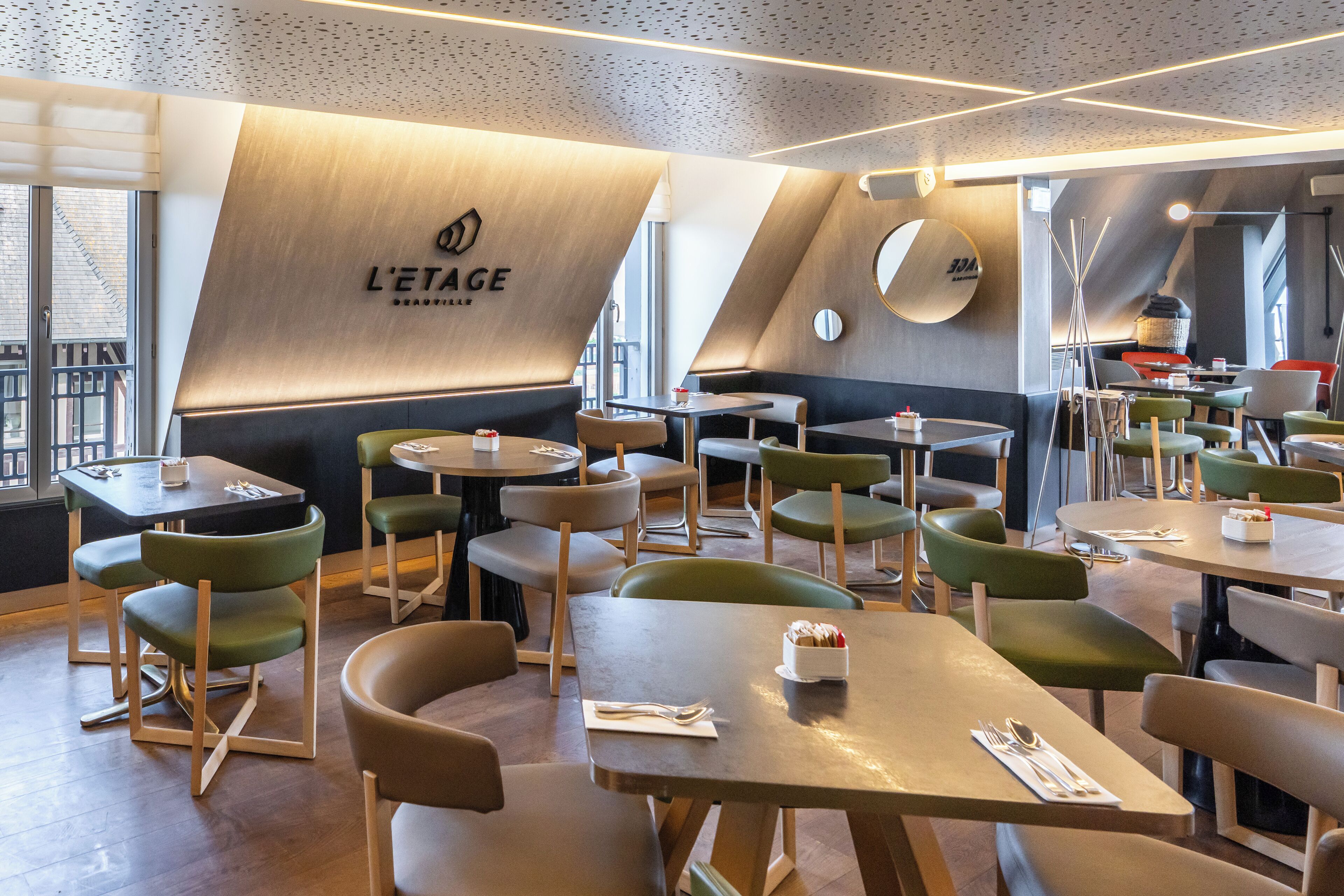A modern restaurant interior with multiple tables and green chairs, a prominent logo on the wall, and a view of the city through large windows.