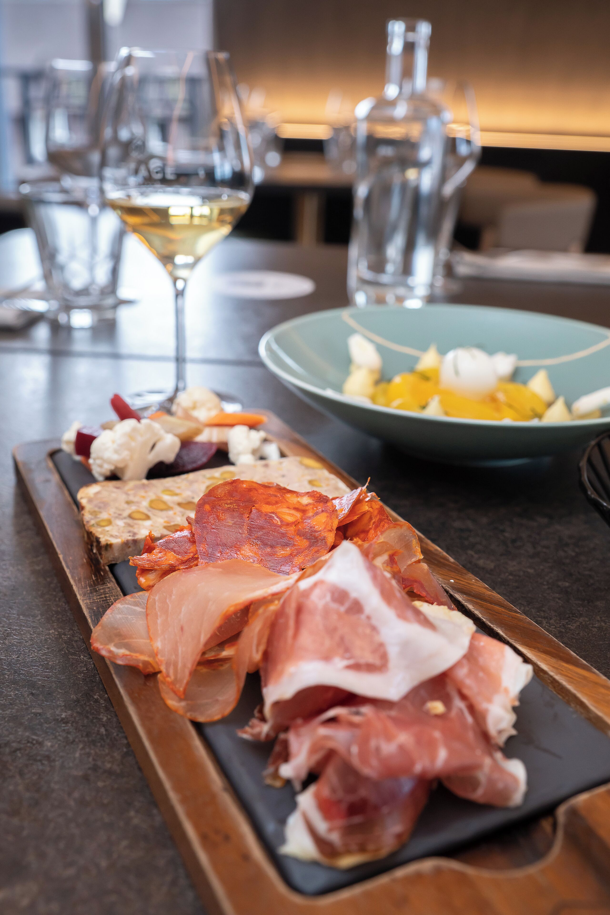 Eine Wurst- und Käseplatte mit verschiedenen Fleisch- und Käsesorten, eine Obstschale und ein Glas Weißwein auf einem Tisch.