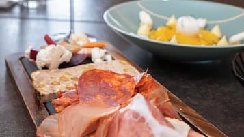 Een charcuterieplank met verschillende vleessoorten en kazen, een schaal fruit en een glas witte wijn op een tafel.