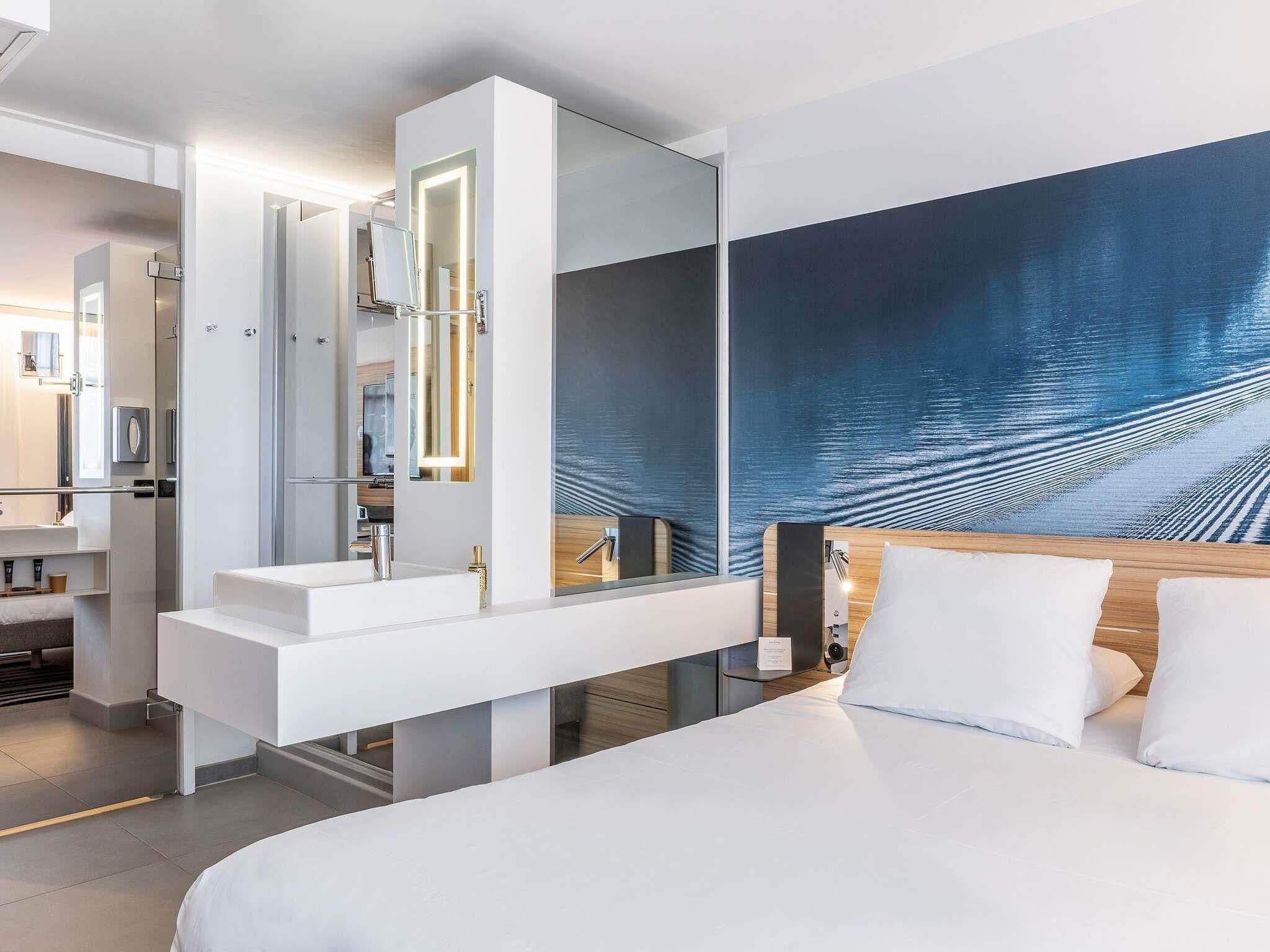 A modern hotel room with a large bed, a built-in vanity with a mirror, a large artwork on the wall, and a view of the sea.