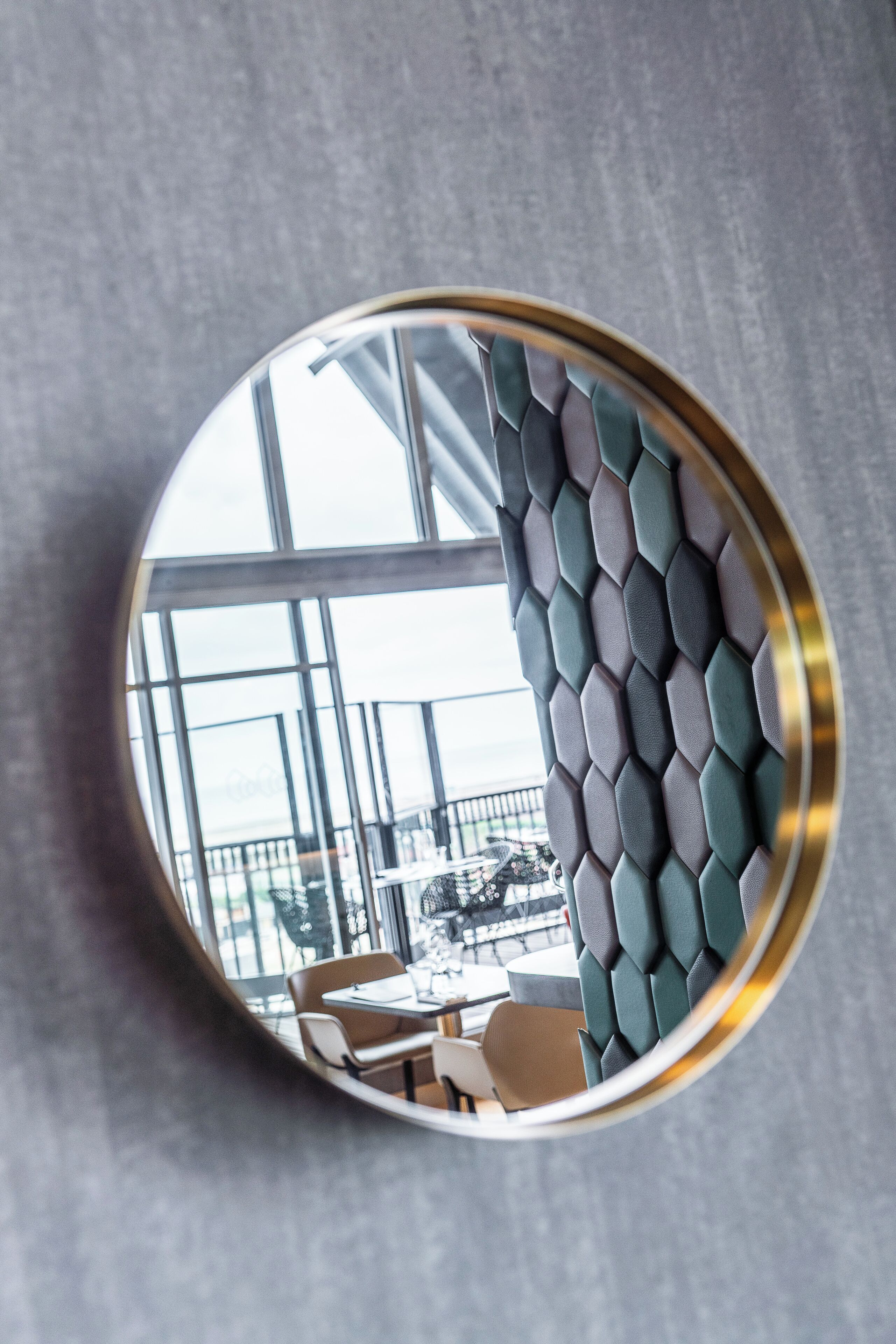 Een ronde spiegel met gouden lijst die een modern interieur weerkaatst, met een geometrisch patroon op de muur en uitzicht op een balkon met zitplaatsen.