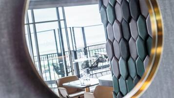 Een ronde spiegel met gouden lijst die een modern interieur weerkaatst, met een geometrisch patroon op de muur en uitzicht op een balkon met zitplaatsen.
