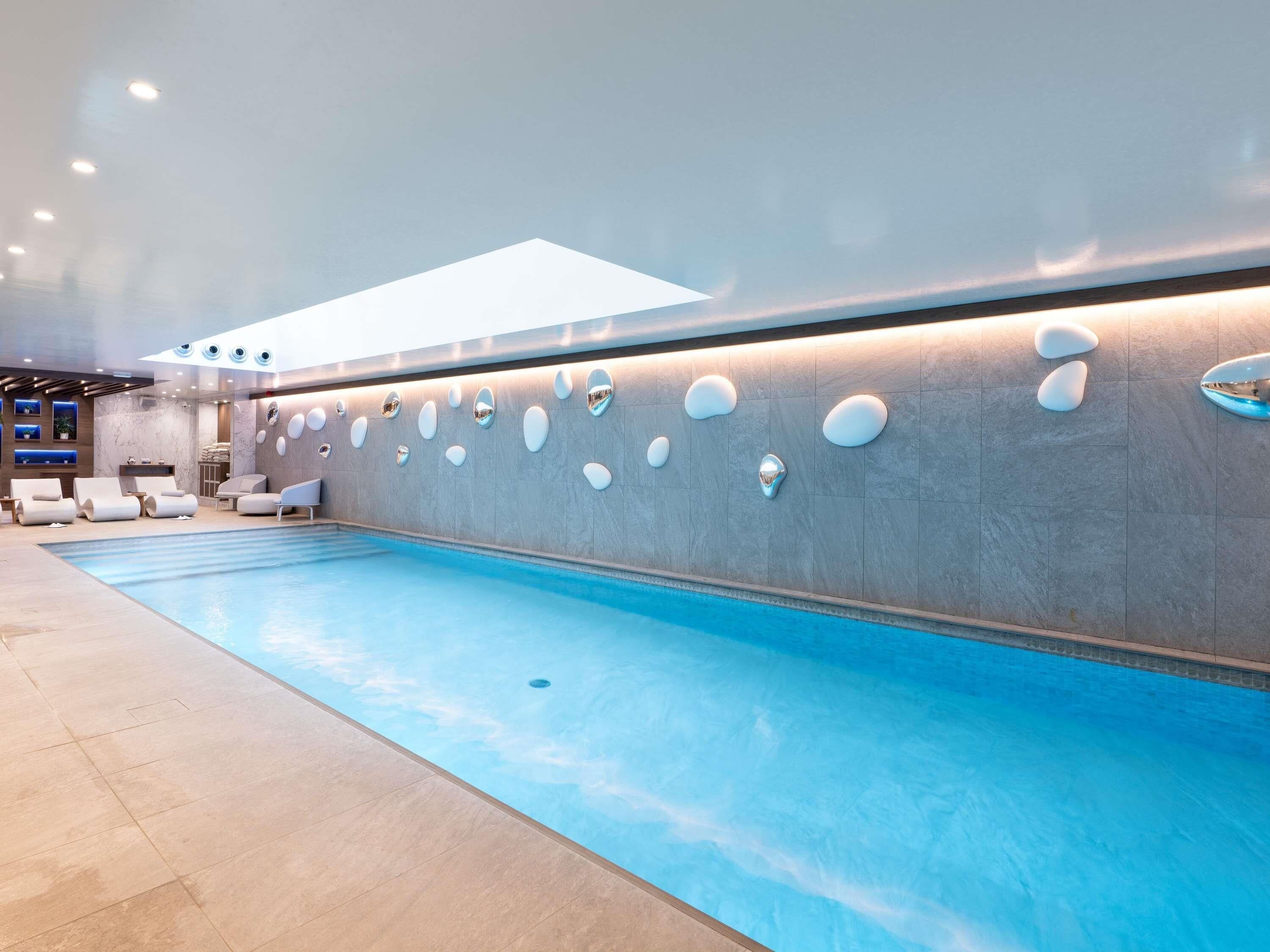 A modern indoor swimming pool with a long, narrow design, surrounded by lounge chairs and a wall with unique, oval-shaped light fixtures.