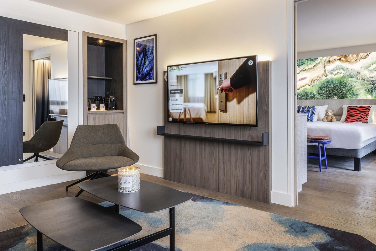 A modern hotel room with a flat-screen TV, a seating area with a table and chairs, a bed with a view, and a mounted artwork.