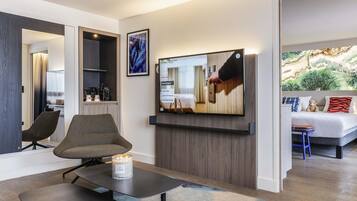 Ett modernt hotellrum med en platt-TV, en sittgrupp med bord och stolar, en säng med utsikt och ett konstverk som hänger på väggen.