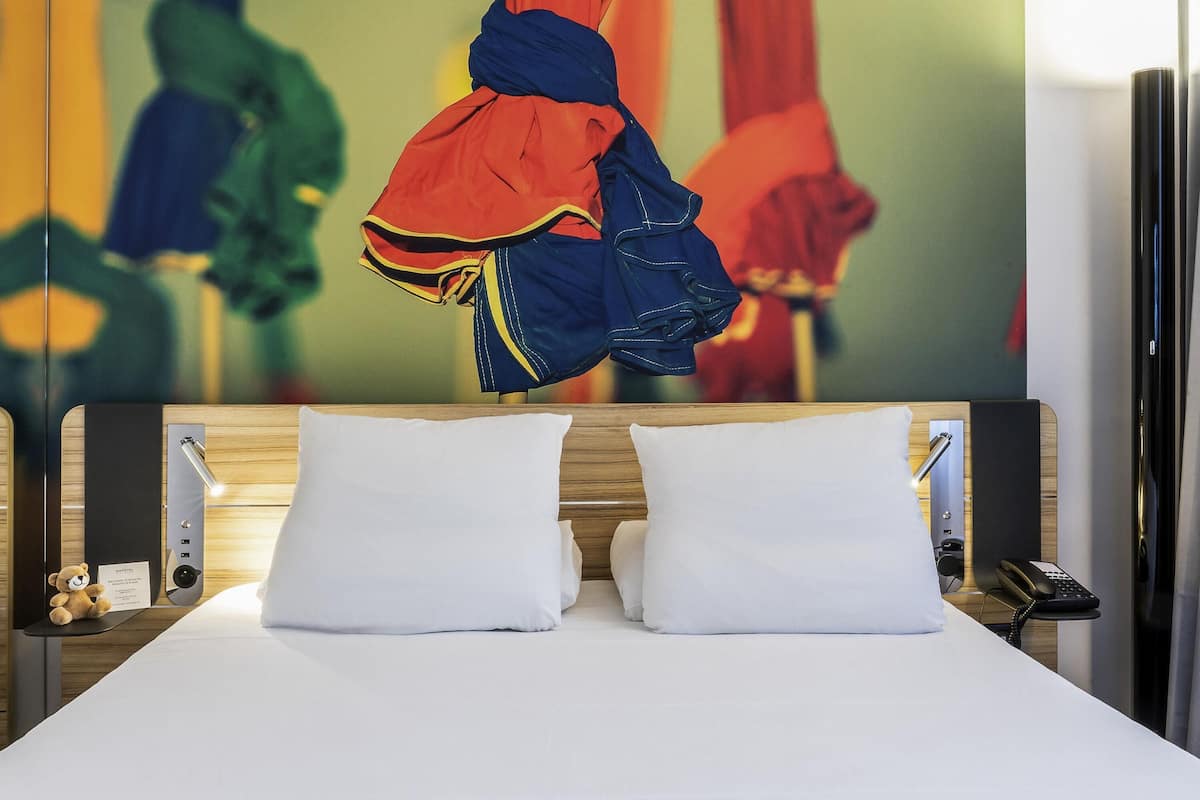 Una habitación de hotel con una cama, almohadas, una mesita de noche con una lámpara, un osito de peluche y una pared decorada con arte abstracto.