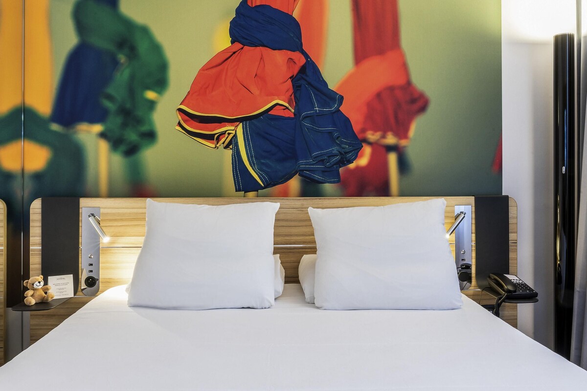 A hotel room with a bed, pillows, a bedside table with a lamp, a teddy bear, and a wall with abstract art.