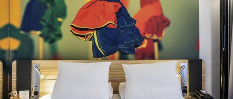 A hotel room with a bed, pillows, a bedside table with a lamp, a teddy bear, and a wall with abstract art.