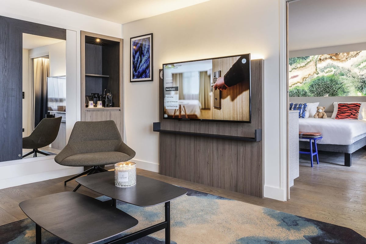 Una habitación de hotel moderna con una televisión de pantalla plana, una zona de estar con una mesa y sillas, una cama con vistas, y una obra de arte colgada en la pared.