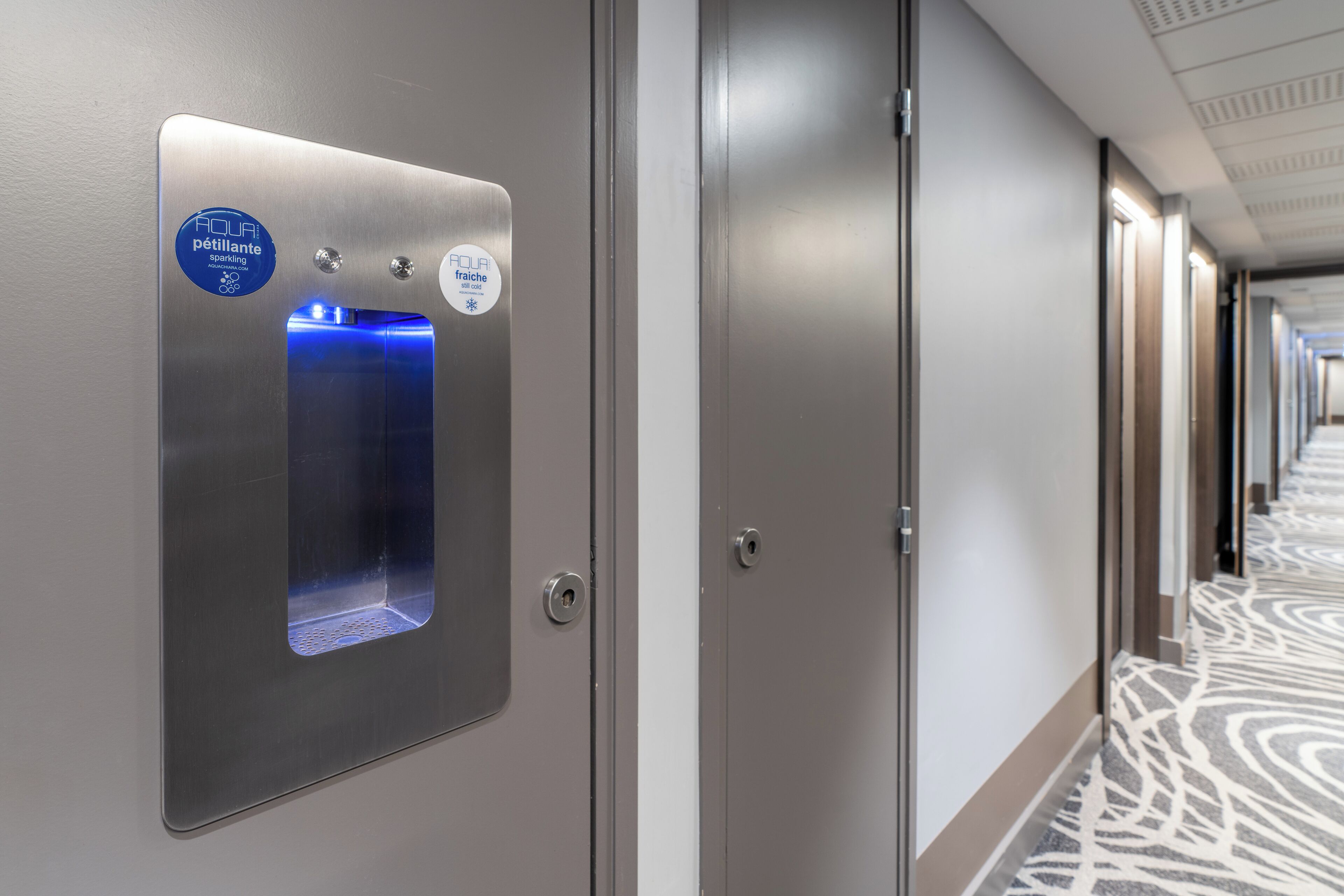 Un couloir d’hôtel avec un distributeur d’eau fixé au mur, une porte et un tapis à motifs.