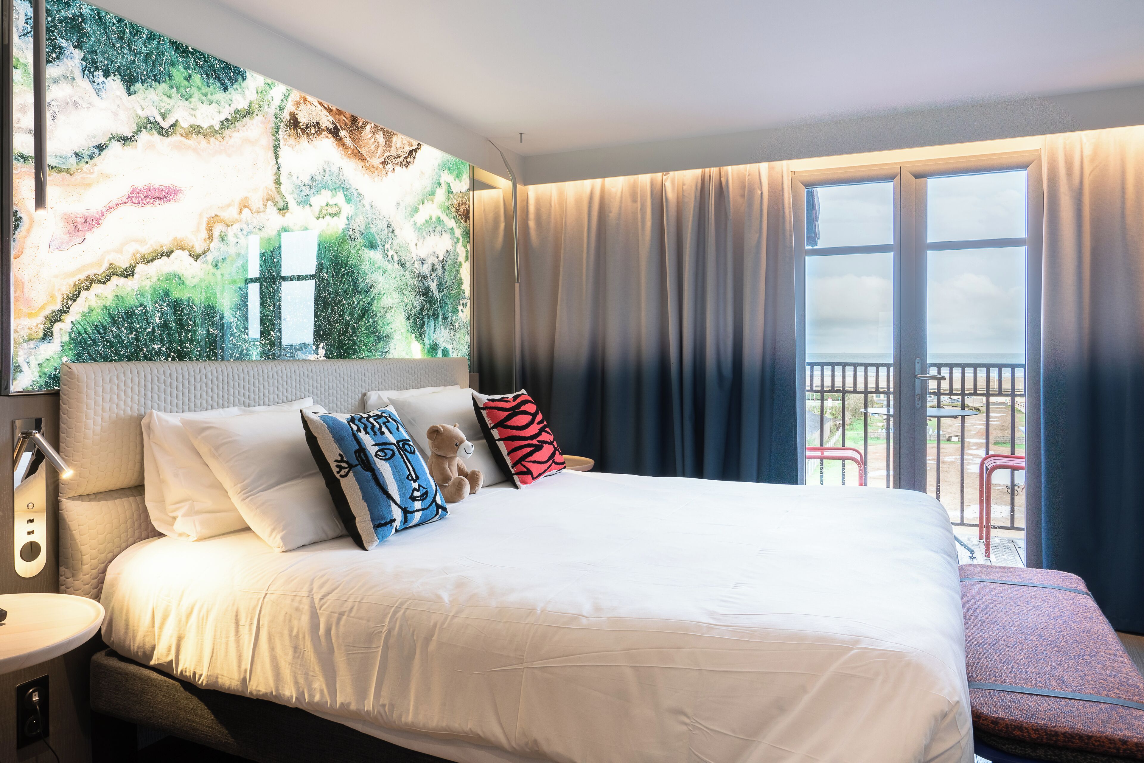A hotel room with a large bed, a nightstand, a wall-mounted lamp, and a balcony with a view.