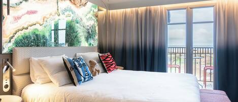 A hotel room with a large bed, a nightstand, a wall-mounted lamp, and a balcony with a view.