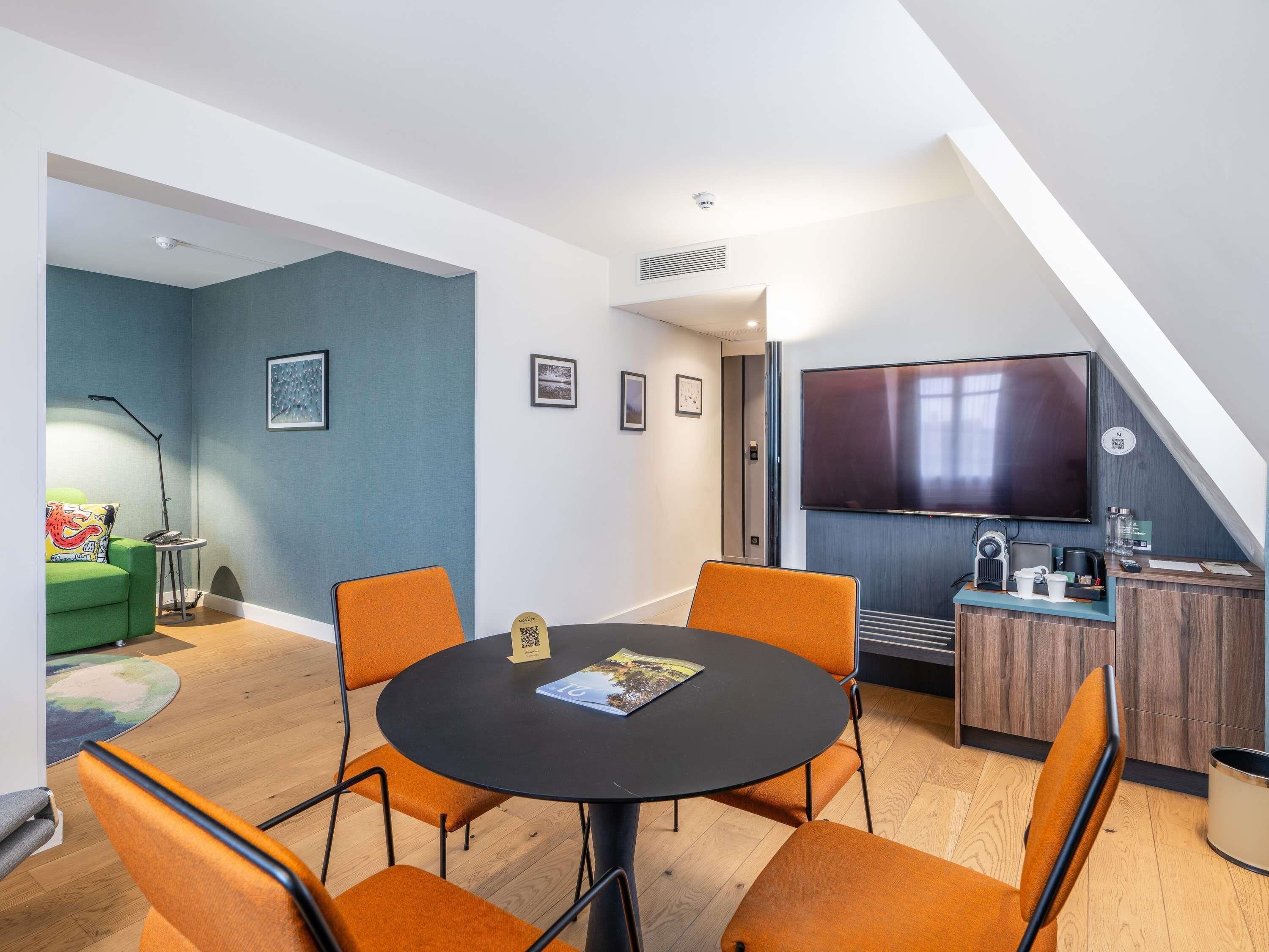 A modern room with a round table, orange chairs, a TV, and a small kitchenette.