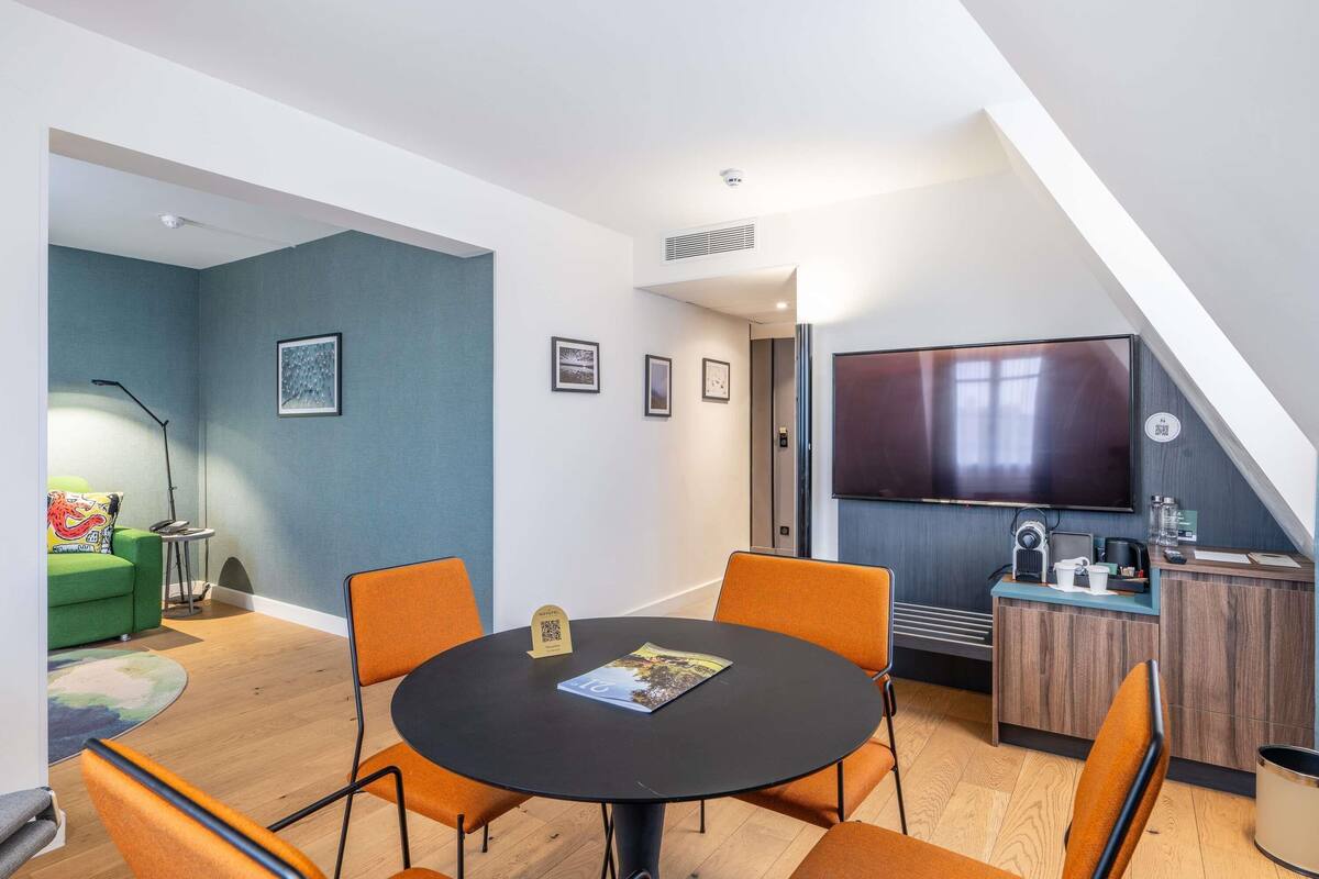 A modern room with a round table, orange chairs, a TV, and a small kitchenette.