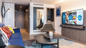 Une chambre d’hôtel moderne équipée d’un canapé, d’une table basse, d’une télévision et d’une étagère murale où sont disposés des articles de toilette.