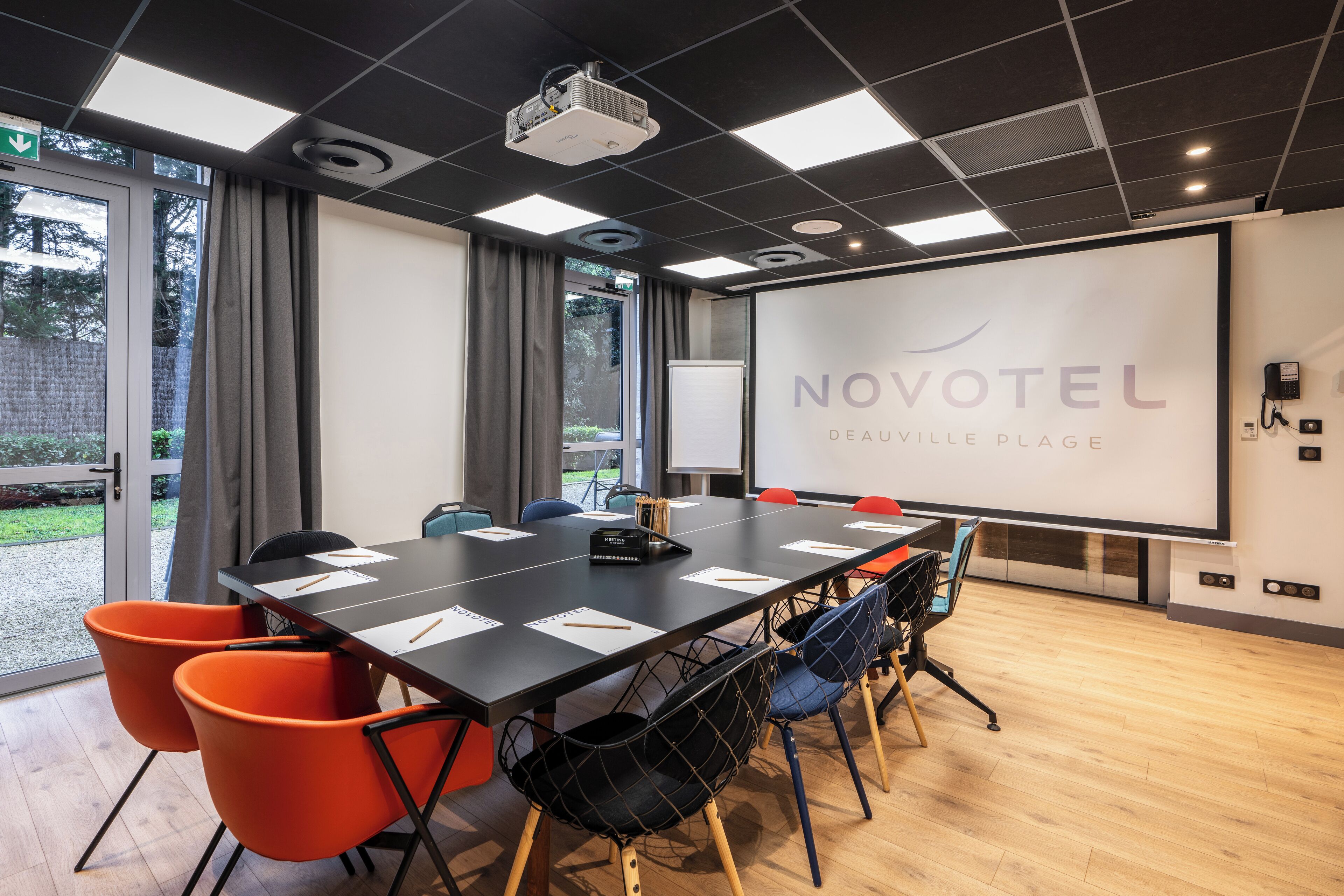 A conference room with a large table, chairs, a projector, and a screen displaying 'NOVOTEL DEAUVILLE PLAGE'.