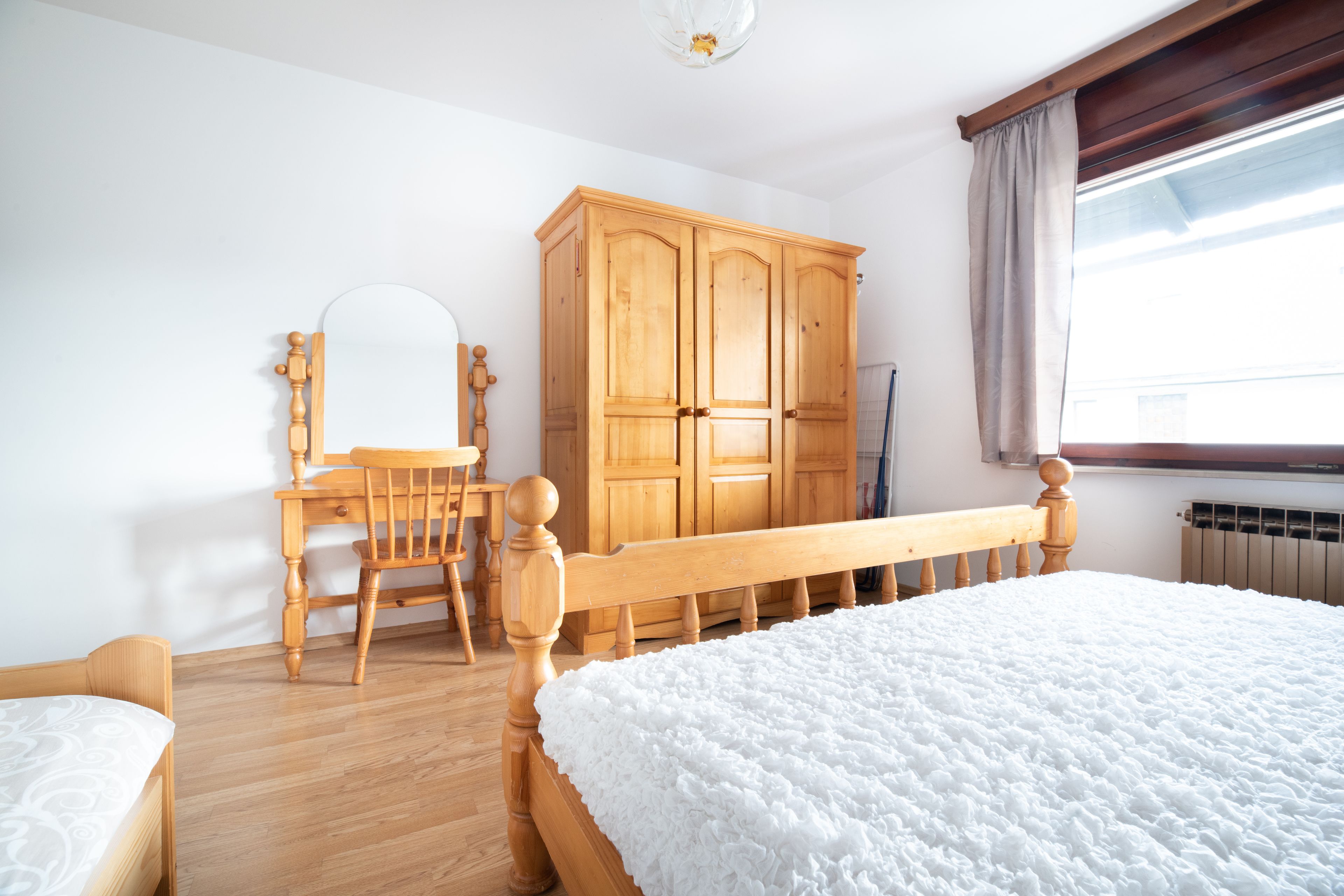 Quarto com cama de madeira, escrivaninha de madeira com cadeira, guarda-roupa e janela com cortinas.