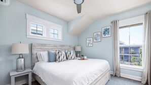 Une chambre à coucher avec un grand lit, deux tables de chevet, un ventilateur de plafond et une fenêtre avec des rideaux.