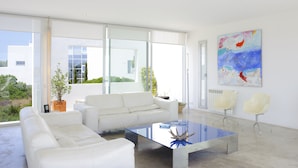 Een moderne woonkamer met een witte bank, een glazen salontafel en twee witte stoelen. Grote ramen bieden uitzicht op het omliggende landschap.