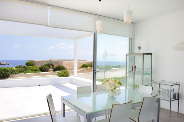 A modern dining area with a glass table and chairs, a vase of flowers, and a view of the sea and landscape.