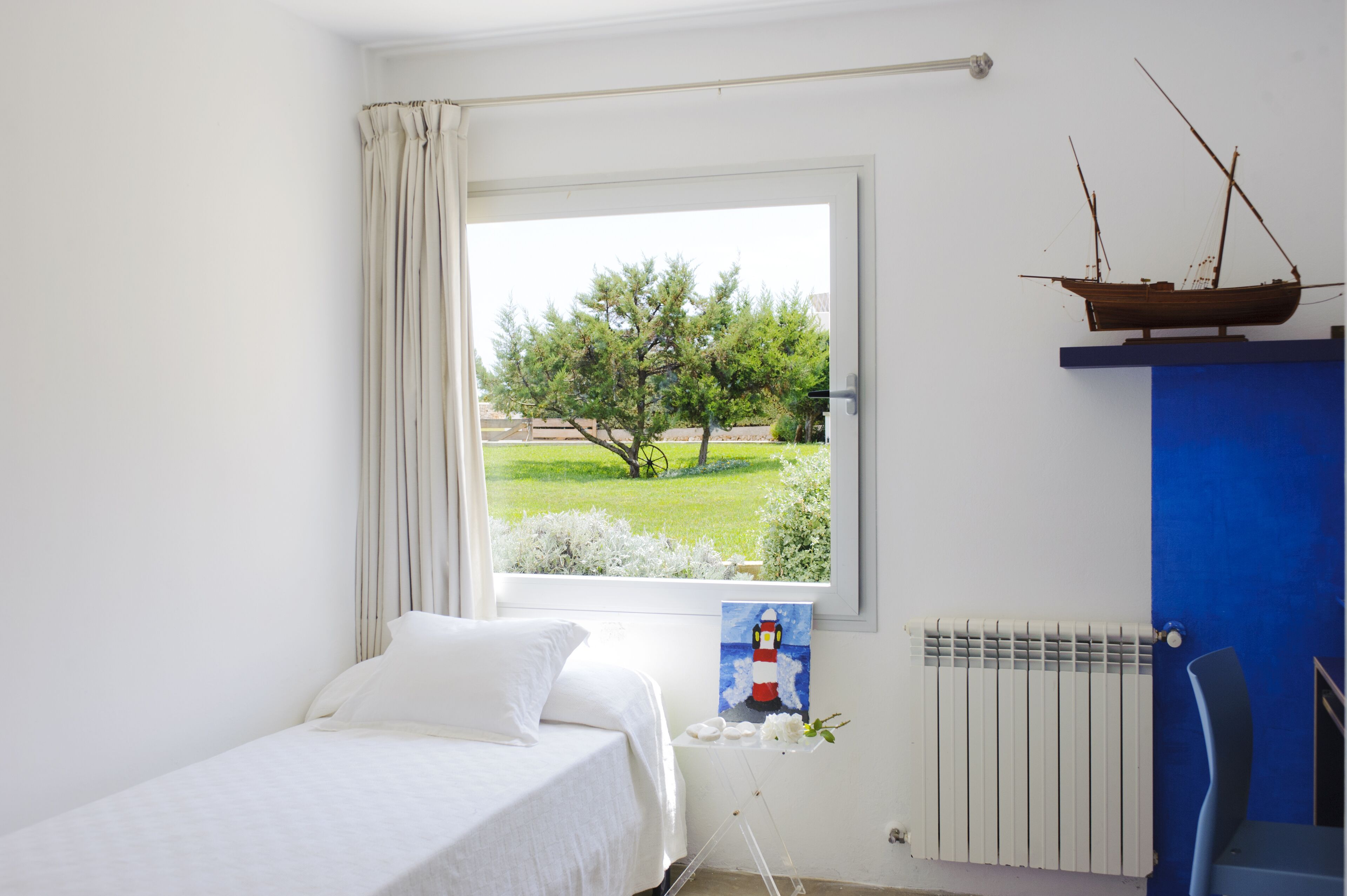 Une chambre à coucher avec un lit, une fenêtre donnant sur un arbre et de l’herbe, un tableau représentant un phare et une maquette de bateau sur une étagère bleue.