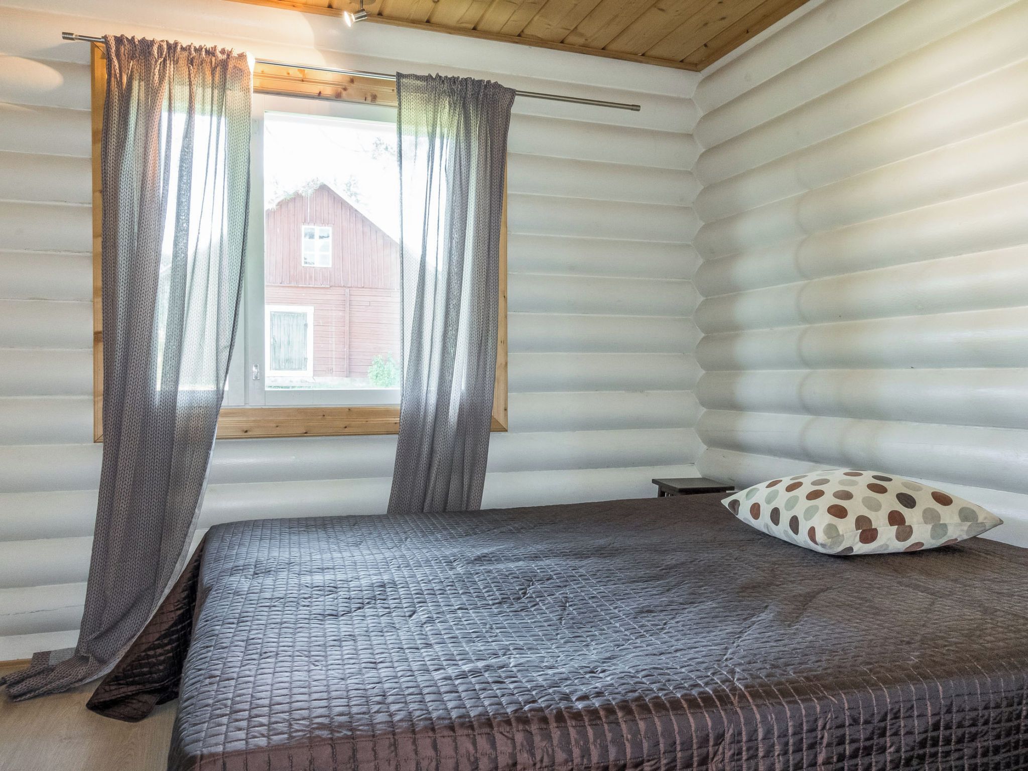 Une chambre avec un lit, une fenêtre avec des rideaux et une vue sur une grange rouge.