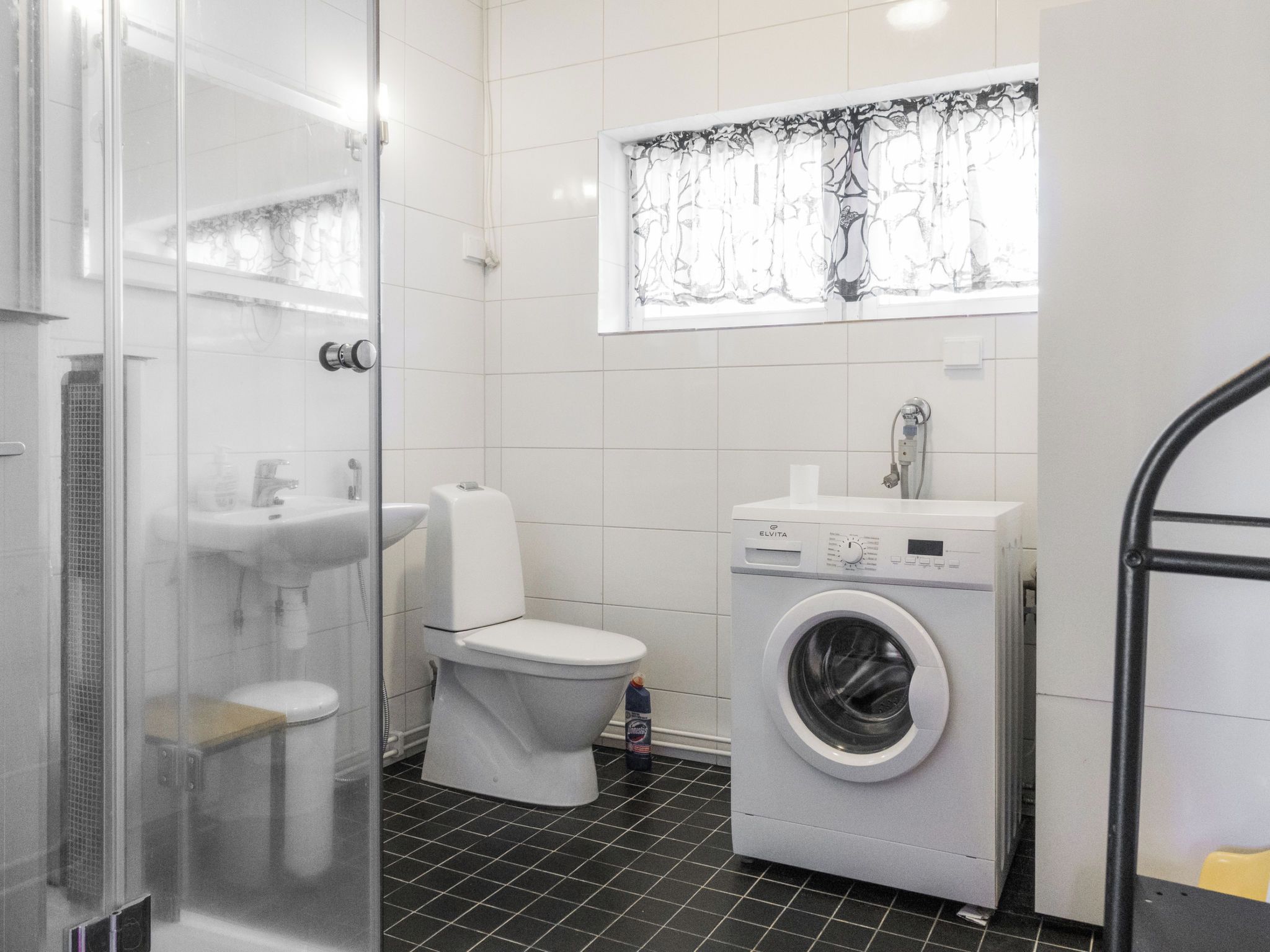 Een moderne badkamer met een glazen douchecabine, toilet, wasmachine en een raam met een decoratief gordijn.