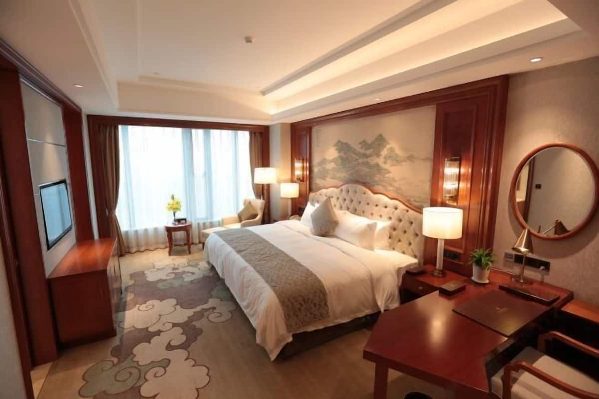 Una habitación de hotel con una cama grande, lámparas de noche, un escritorio, una silla, un televisor y un ventanal con cortinas.