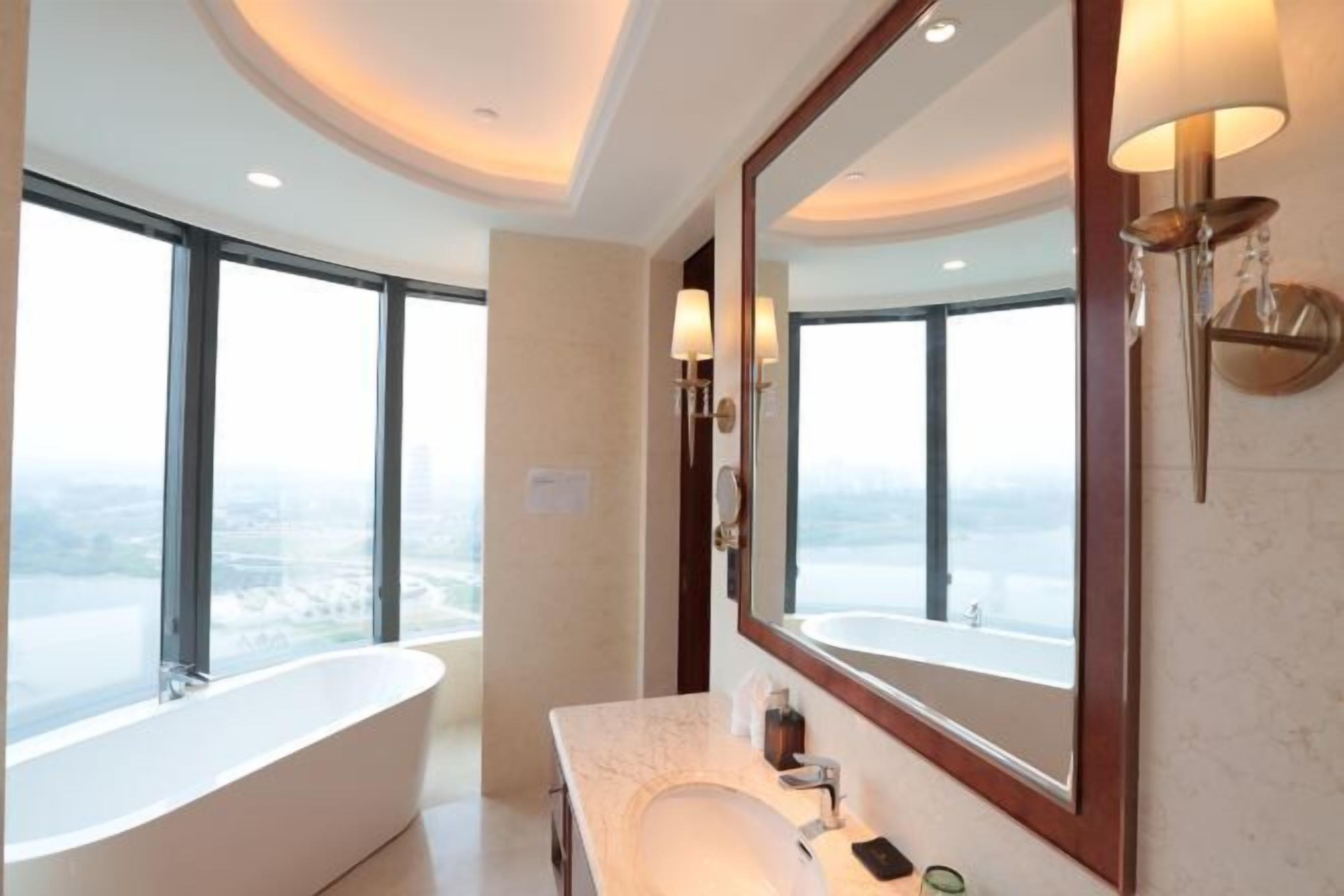 Un baño moderno con una bañera grande, un tocador con espejo y vistas a la ciudad a través de amplias ventanas.