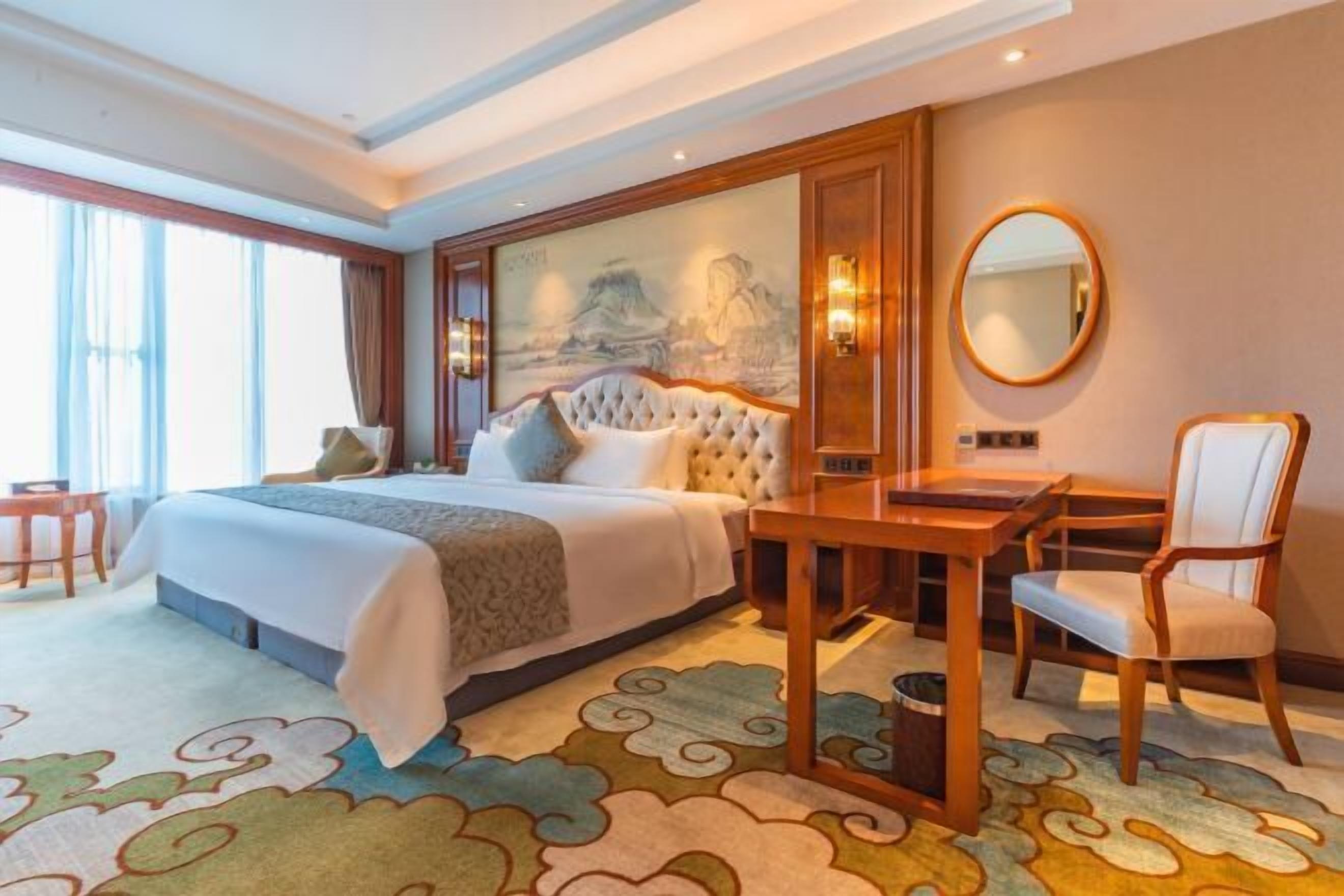 A hotel room with a large bed, a desk, a chair, a round mirror, and a painting on the wall.