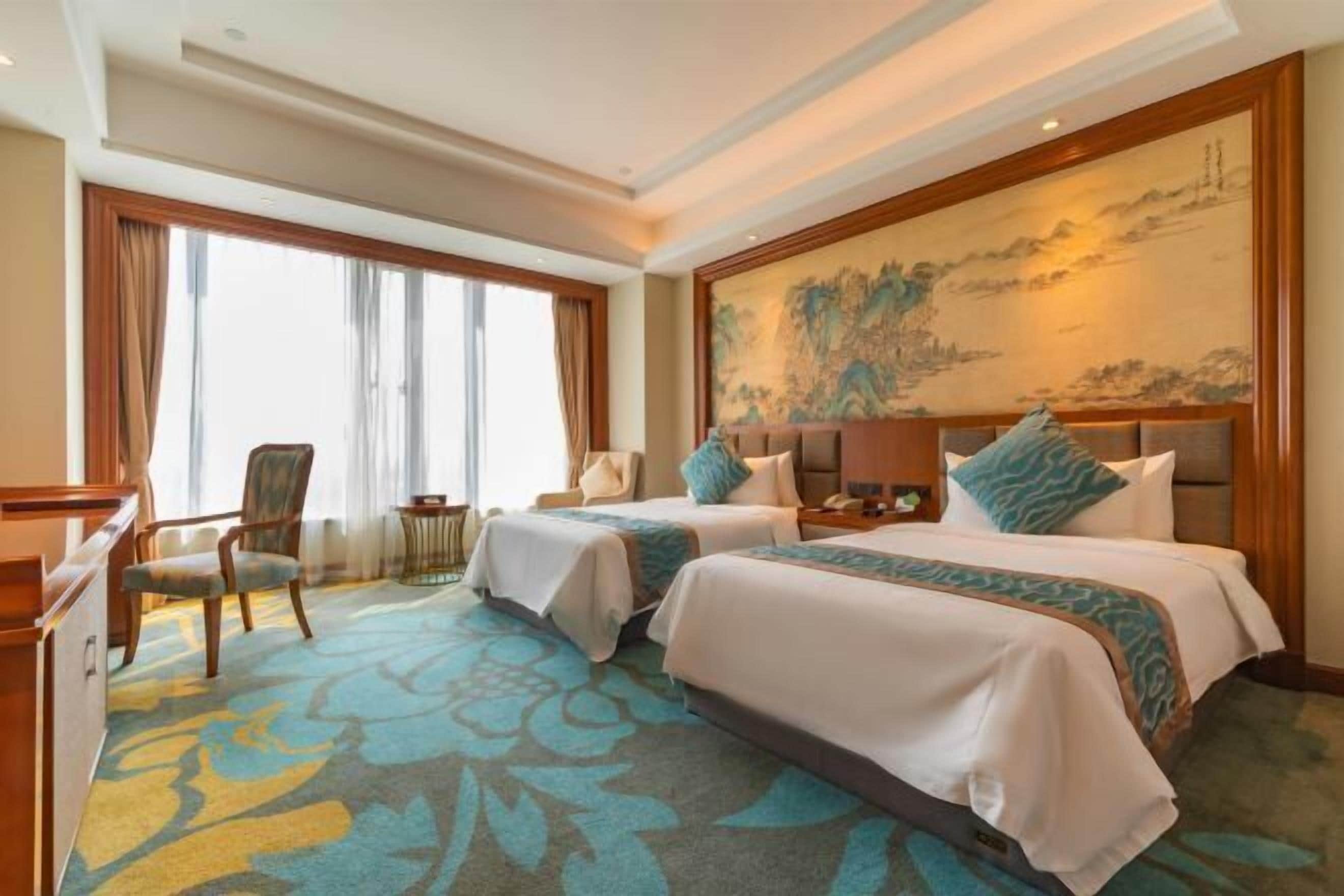 Une chambre d’hôtel avec deux lits, un bureau et une chaise. Il y a un grand tableau au mur et une fenêtre avec des rideaux.