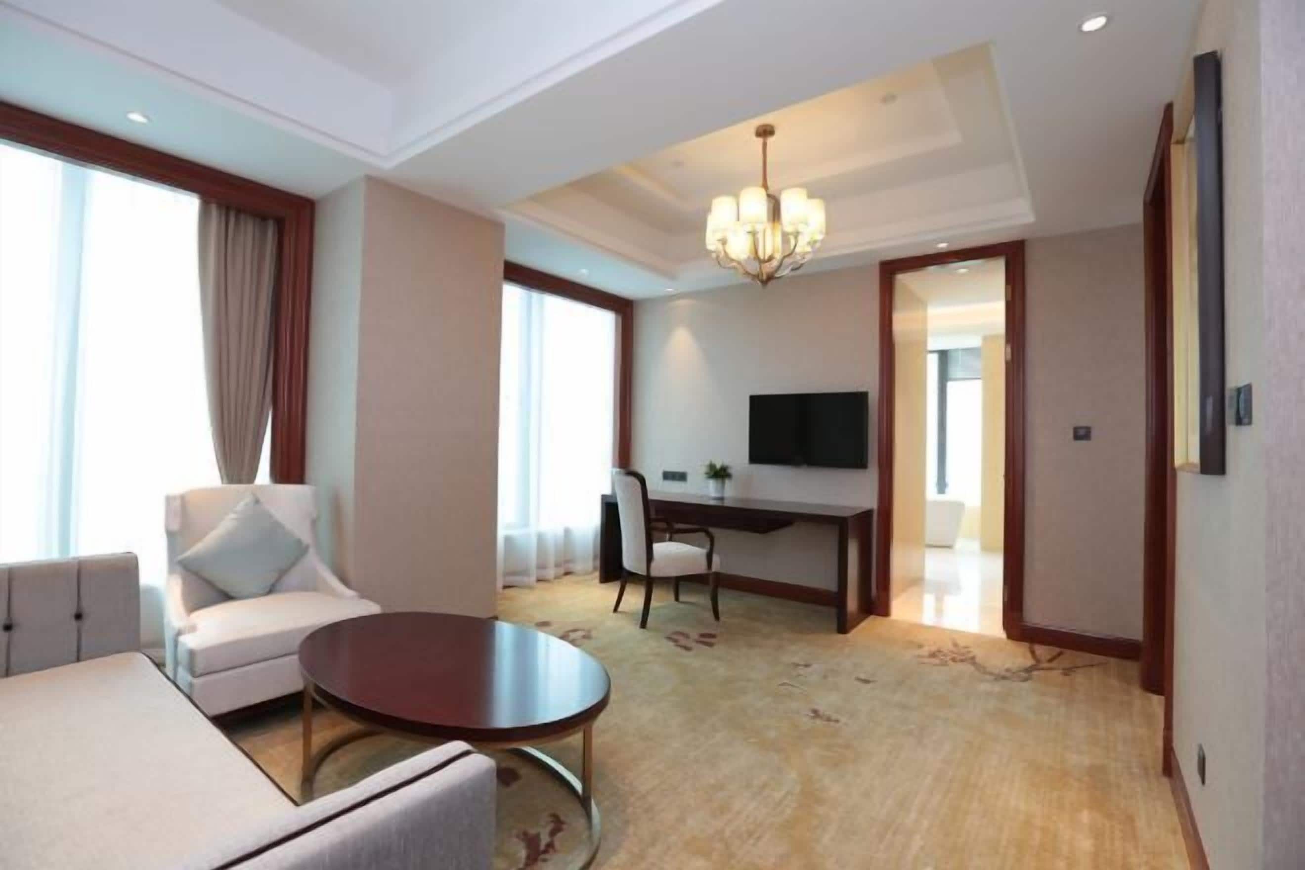 A hotel room with a sofa, a round table, a desk with a chair, a TV, and a chandelier.