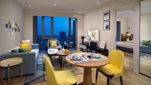 A modern hotel room with a sofa, dining table, chairs, a bed, a TV, and a city view.