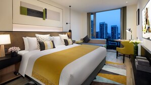 A hotel room with a large bed, a desk, a chair, a view of the cityscape, and a city skyline visible through the window.