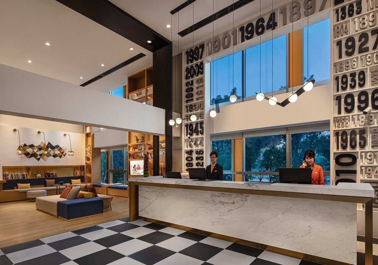 A modern hotel lobby with a checkered floor, a reception desk, and a seating area with a sofa and bookshelves.