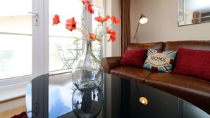 Una sala de estar con un jarrón de vidrio sobre una mesa, un sofá de cuero marrón con un cojín rojo, y un espejo en la pared.