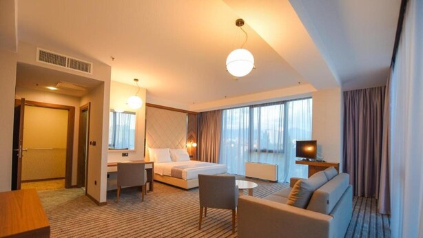 Une chambre d’hôtel avec un lit, un bureau, un canapé, une télévision et de grandes fenêtres.