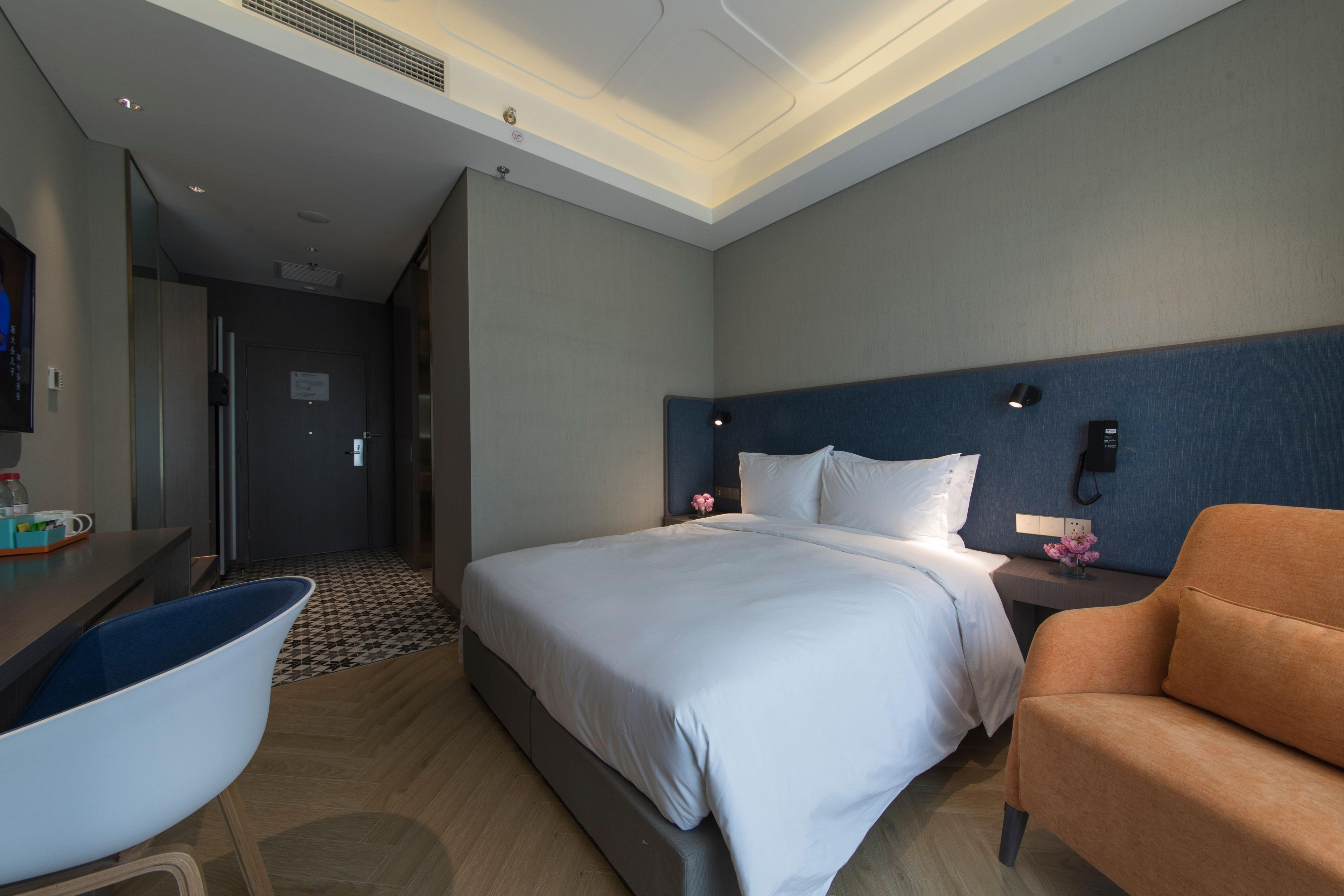 A hotel room with a bed, a desk, a chair, and a nightstand with a lamp.