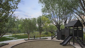 A playground with a slide and climbing structures, surrounded by trees and a paved path.