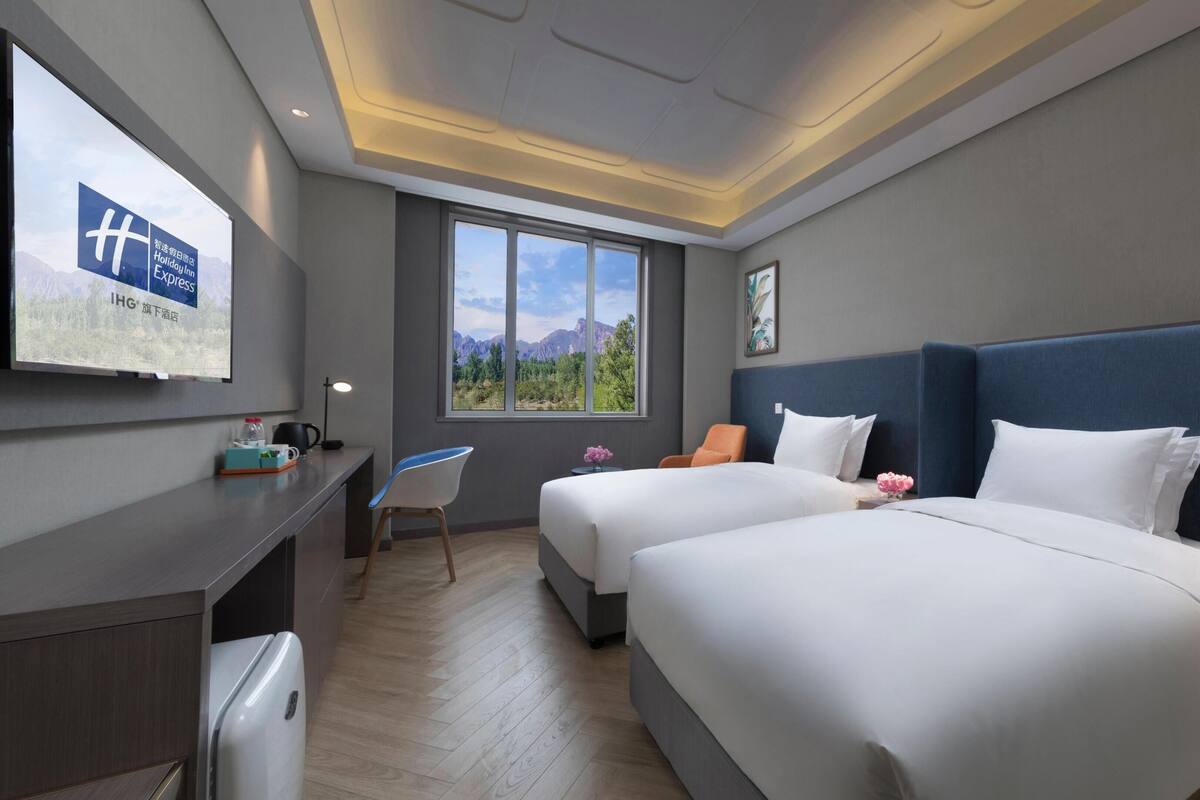 Ein Hotelzimmer mit zwei Betten, einem Schreibtisch, einem Stuhl, einem Fenster mit Bergblick und einem an der Wand befestigten Fernseher.