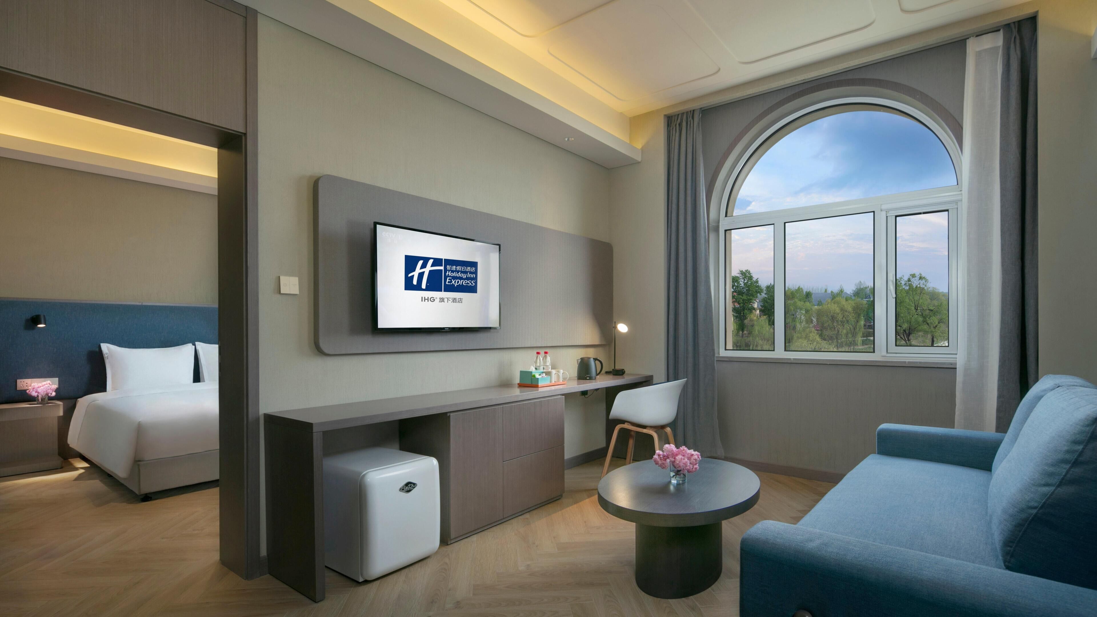 A hotel room with a bed, a desk, a chair, a sofa, a TV, and a large window.