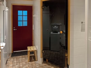 A narrow hallway with a red door featuring a window, a small wooden stool, and a tiled floor.