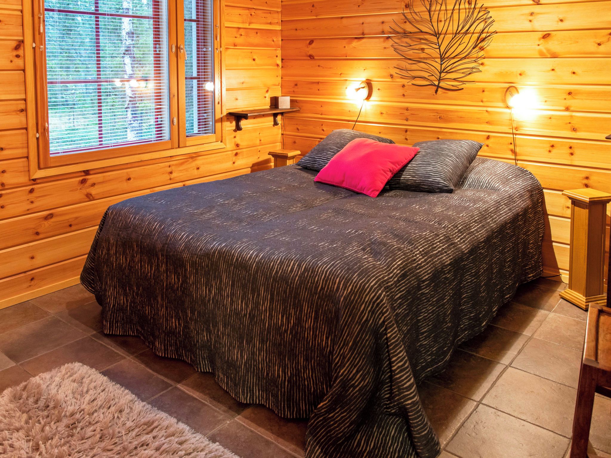 A wooden cabin room with a bed, a window with blinds, a wall-mounted light, and a decorative branch.