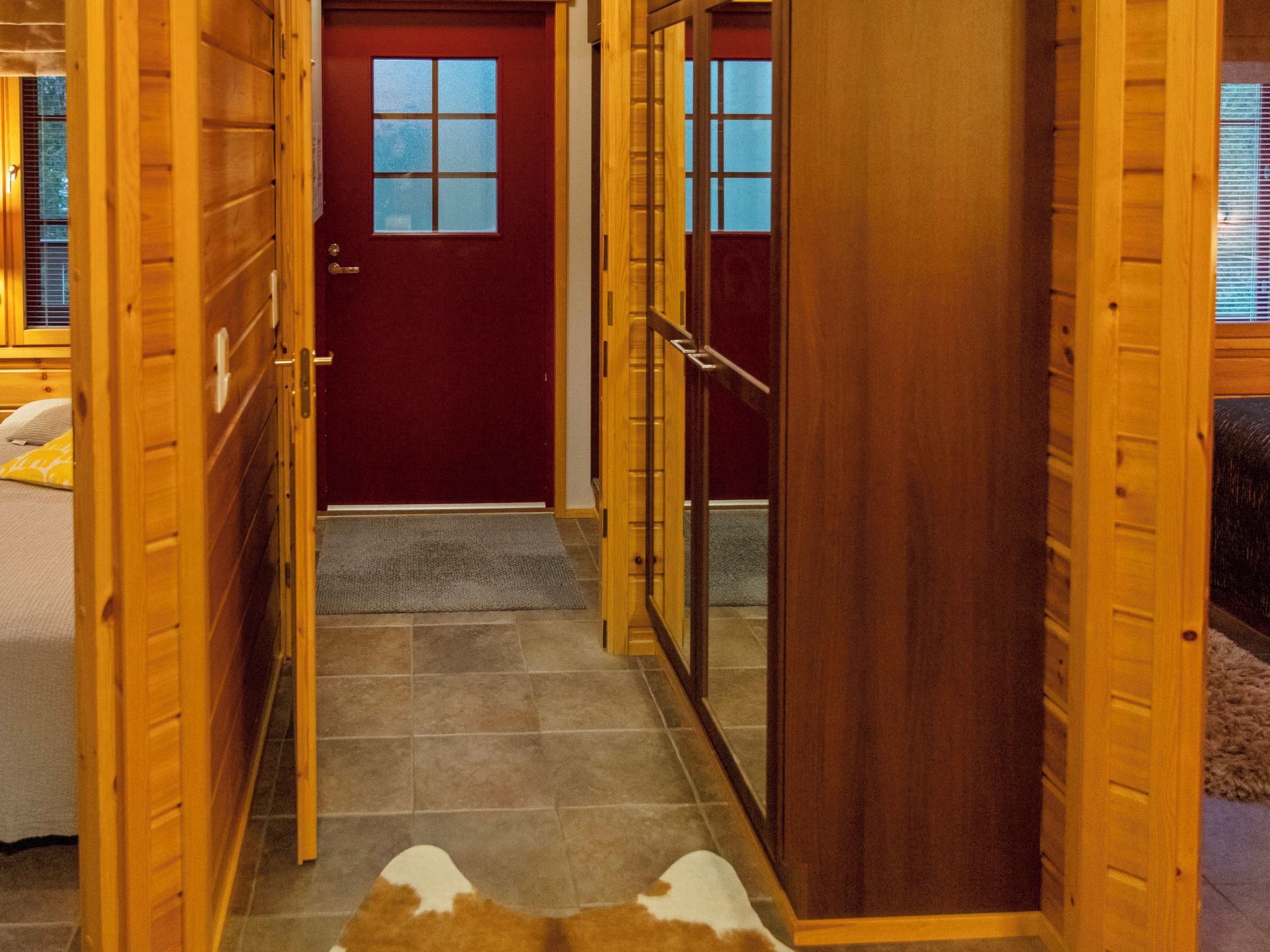 A wooden interior hallway leading to a red door with a window, a wooden door with a handle, and a rug on the floor.