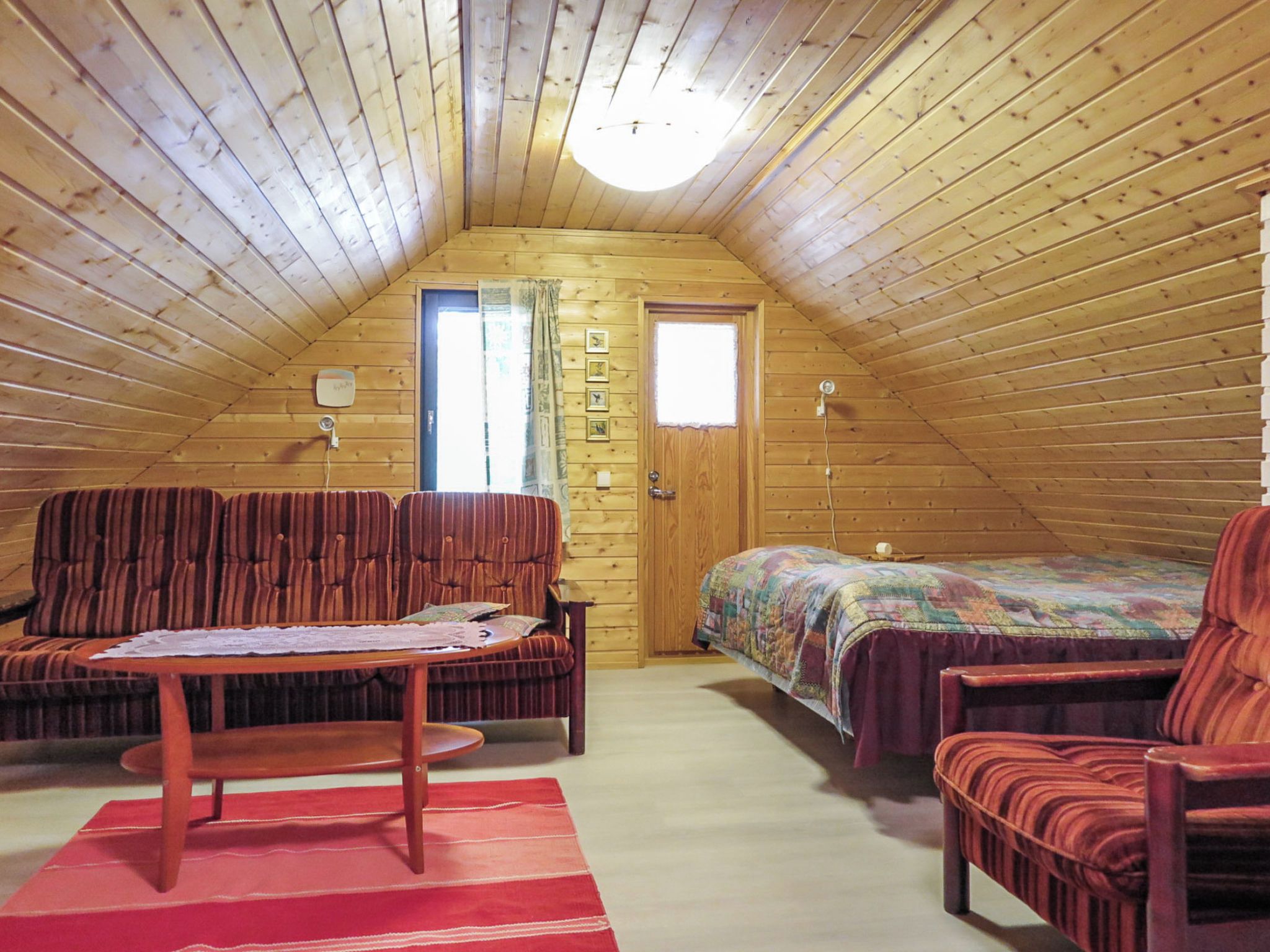 Une chambre mansardée chaleureuse avec un plafond en pente, des murs en bois, un lit simple, deux chaises, une petite table et une fenêtre.
