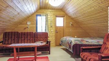 Une chambre mansardée chaleureuse avec un plafond en pente, des murs en bois, un lit simple, deux chaises, une petite table et une fenêtre.