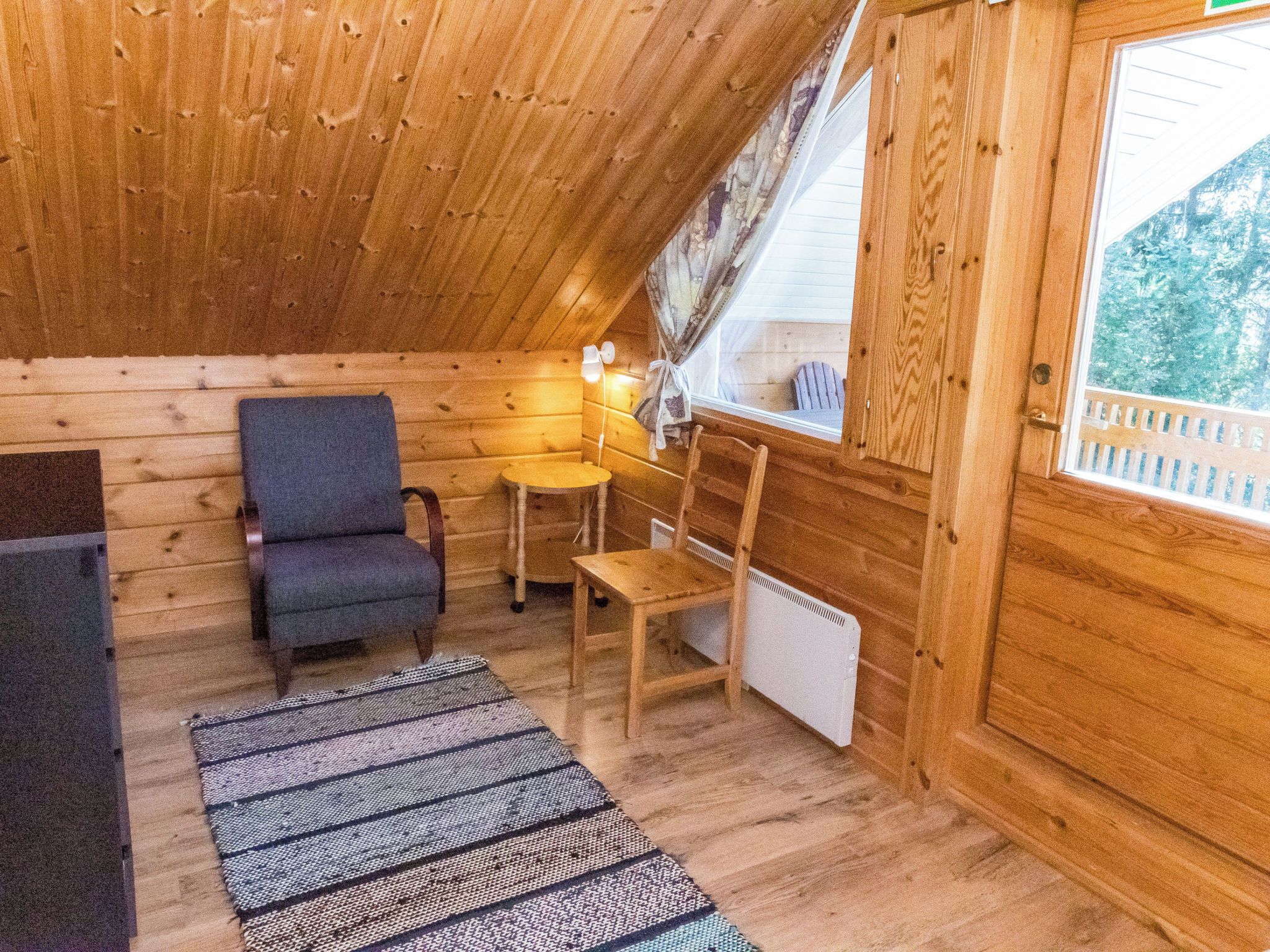 A cozy cabin interior with wooden walls, a gray chair, a small table, a radiator, and a window with a view of trees.