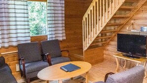 Interno di una baita in legno con una scala, due poltrone, un tavolo rotondo, un televisore e una finestra con tende.